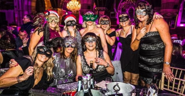 9 women in festive masks and dresses at a masquerade Christmas party, smiling joyously around a table with drinks