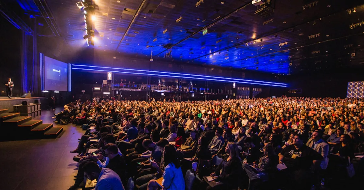 A packed conference full of an attentive audience facing a stage where a speaker is presenting