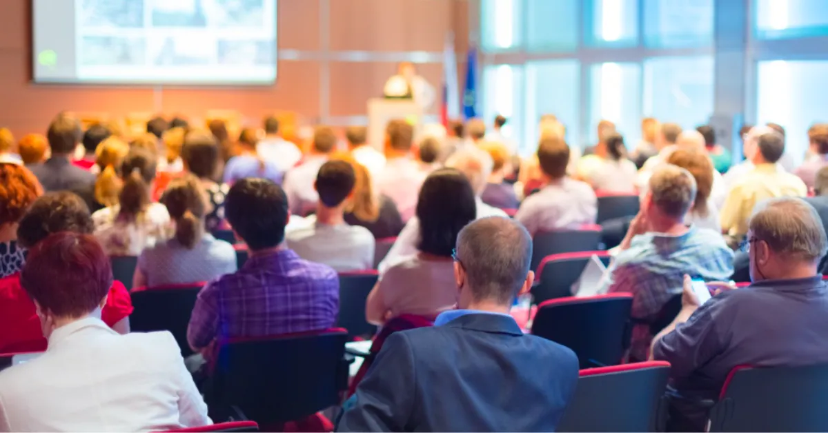 A diverse audience sits in a conference room, attentively facing a speaker at a podium
