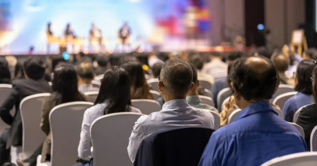 Conference audience seated indoors, facing a stage with blurred figures of a panel