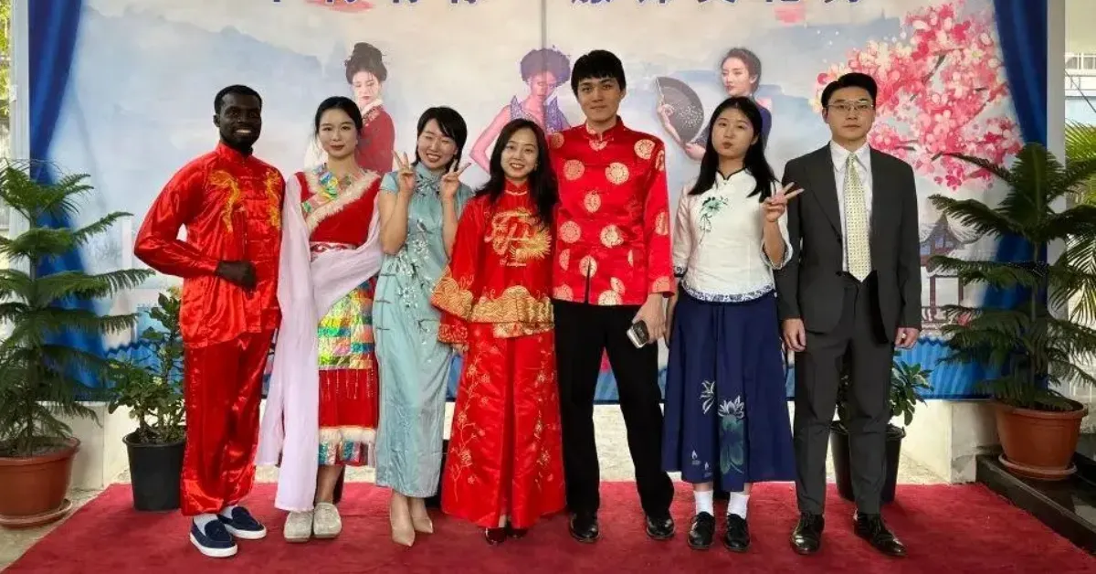 A diverse group of seven people stands on a red carpet, wearing colorful, traditional Chinese New Year attire
