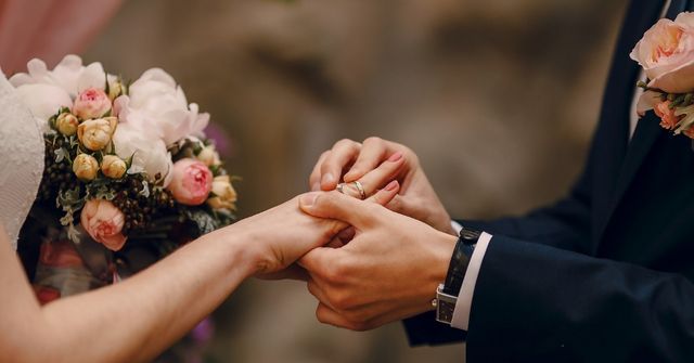 A groom places a ring on the bride's finger, against a blurred background