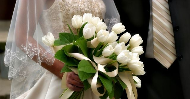Bride holding a bouquet of white tulips with green leaves and stands beside a groom