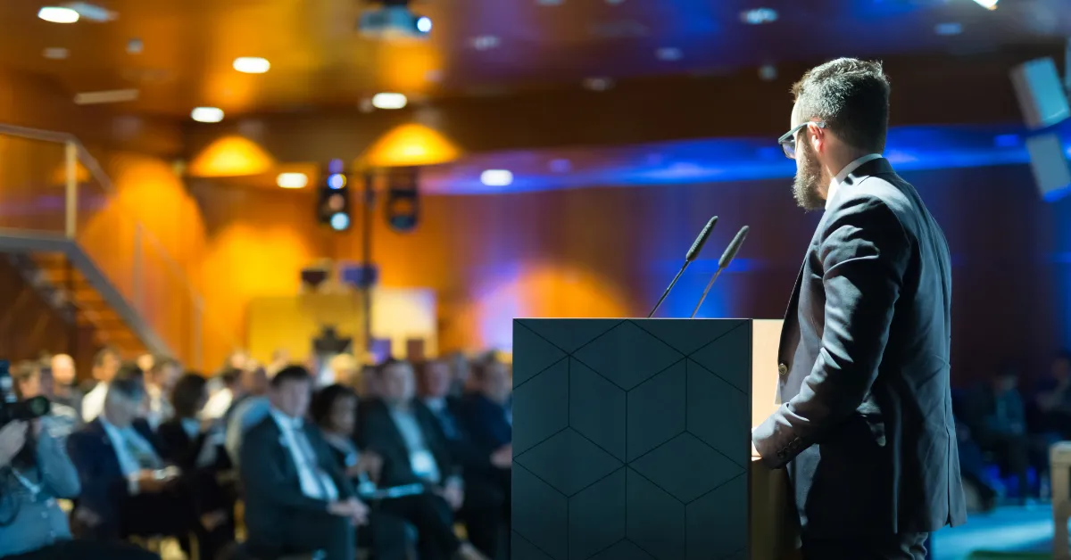 A man in a suit speaks at a podium to a dimly lit audience in a conference room