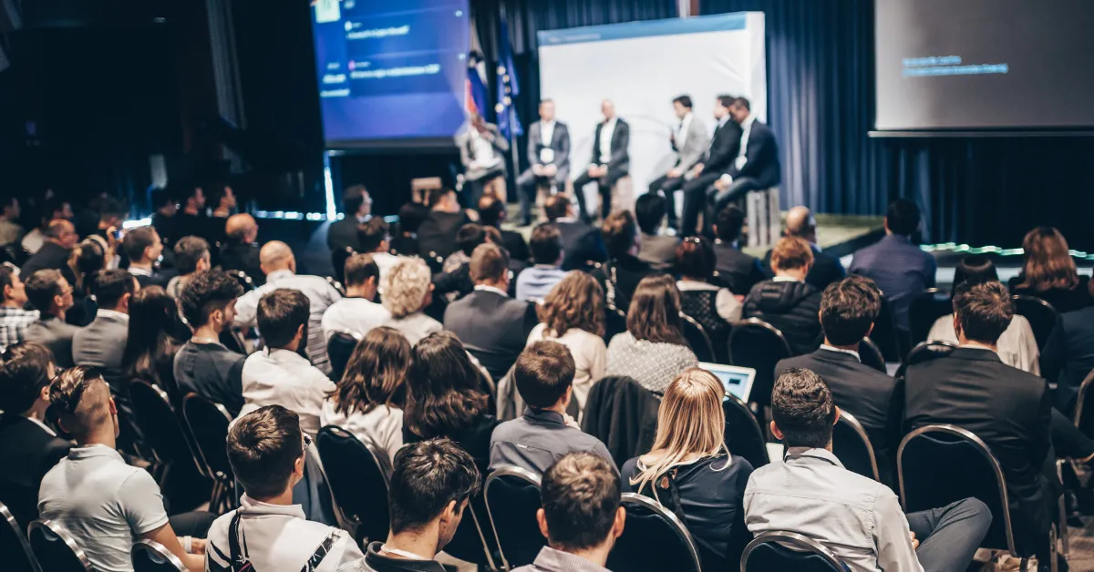 Audience attentively listens to a panel of six speakers on stage at a corporate event