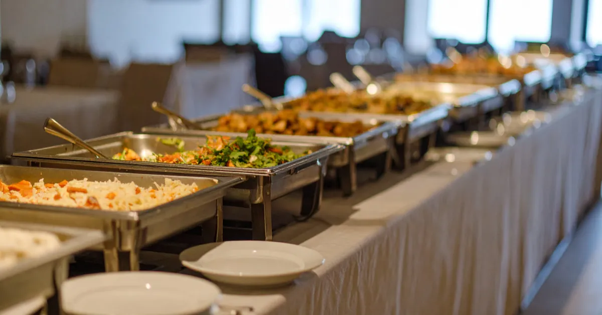 A long table with a white cloth holds a buffet with several metal trays of diverse foods, at a corporate event