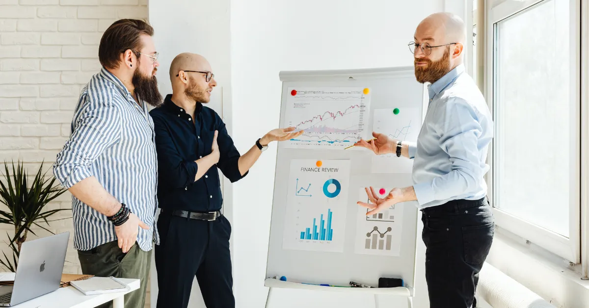 Three colleagues stand around a flipchart with financial graphs discussing data