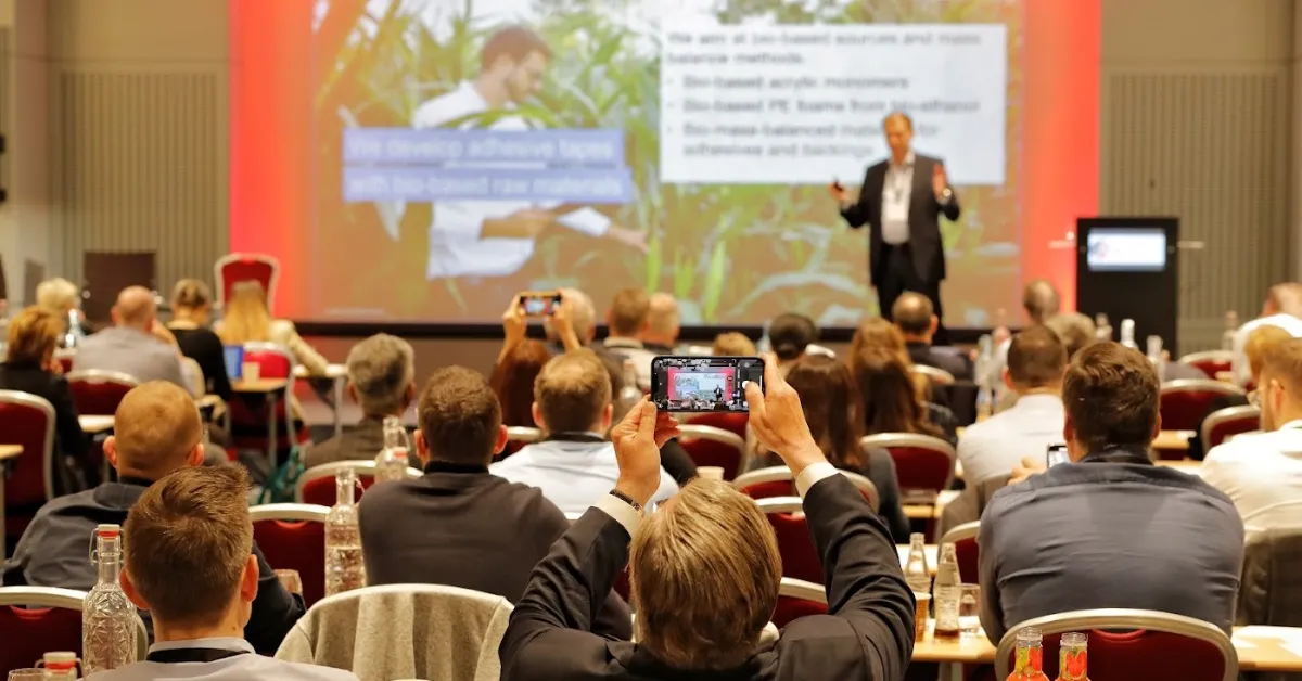 Audience at a seminar listens to a speaker on stage while capturing the presentation with their smartphones