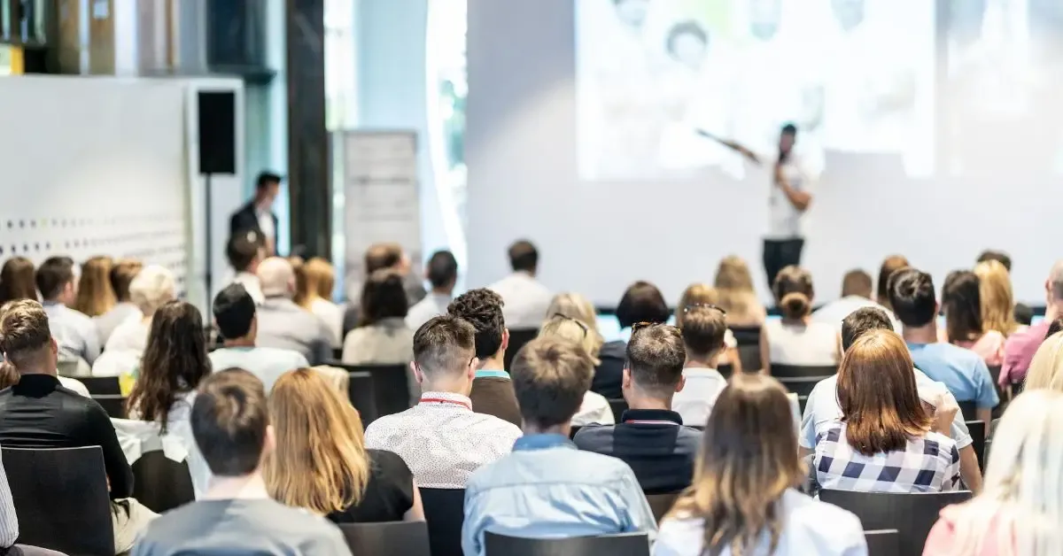 An audience sits in rows, attentively facing a speaker presenting slides at a seminar